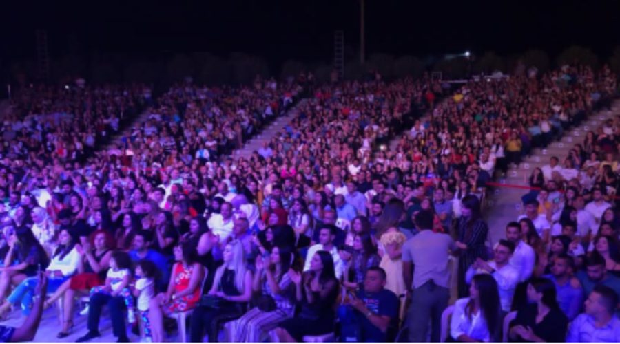 A packed amphitheatre crowd under purple stage lighting